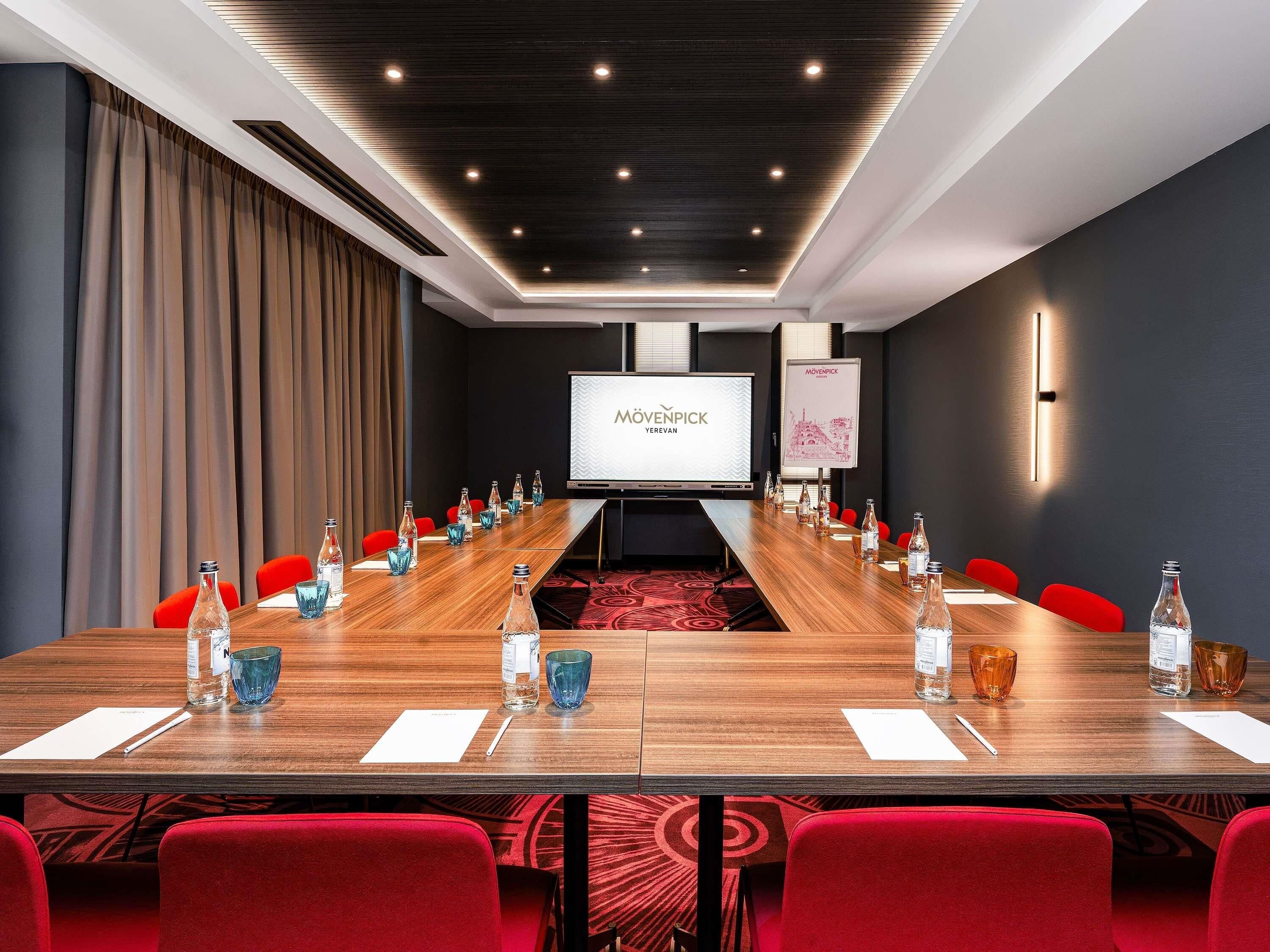 A conference room with a long table, red chairs, and a projector screen displaying 'MÖVENPICK YEREVAN'.