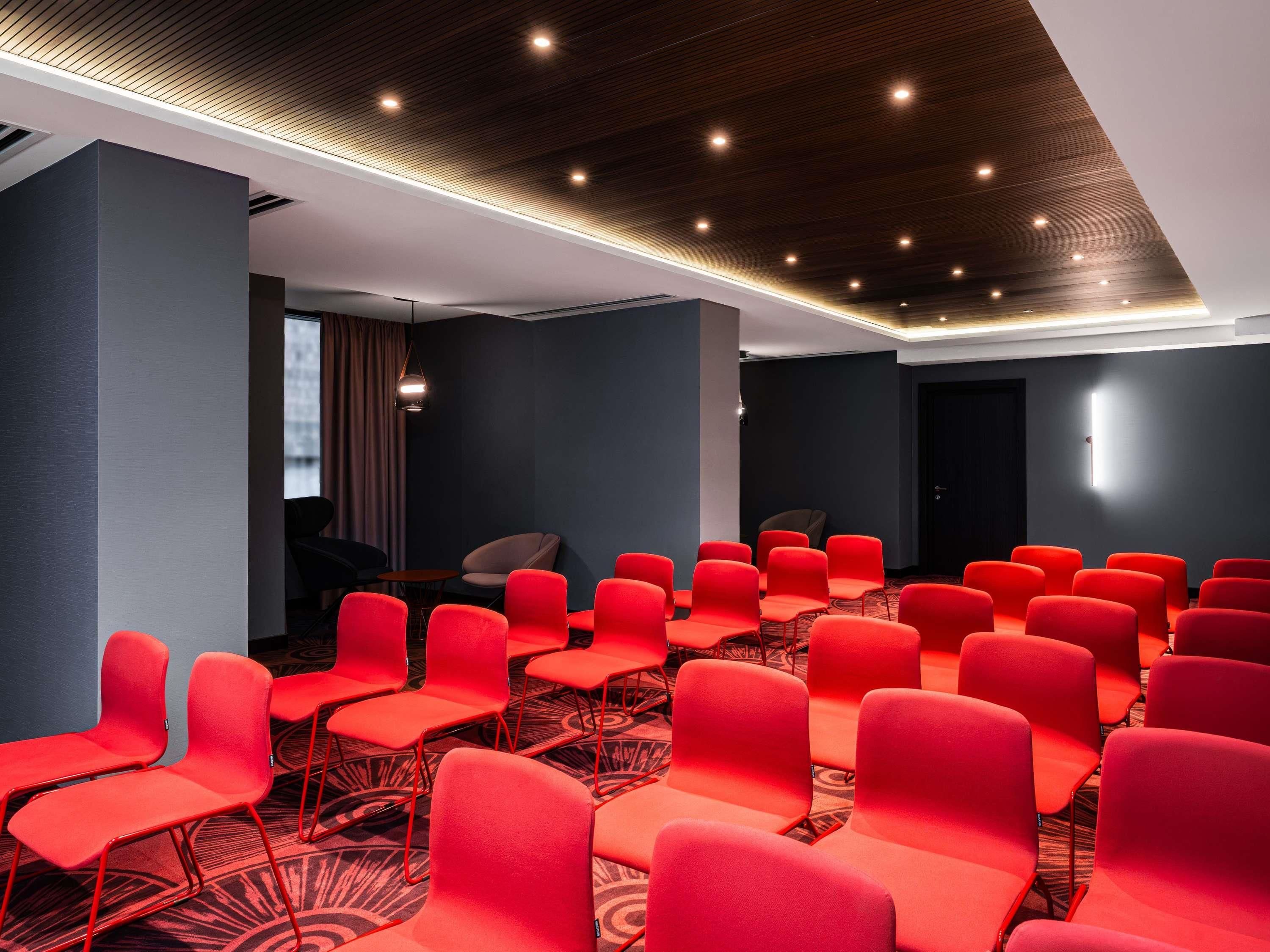 Un salón de conferencias con hileras de sillas rojas, alfombra estampada y un techo oscuro con iluminación empotrada.