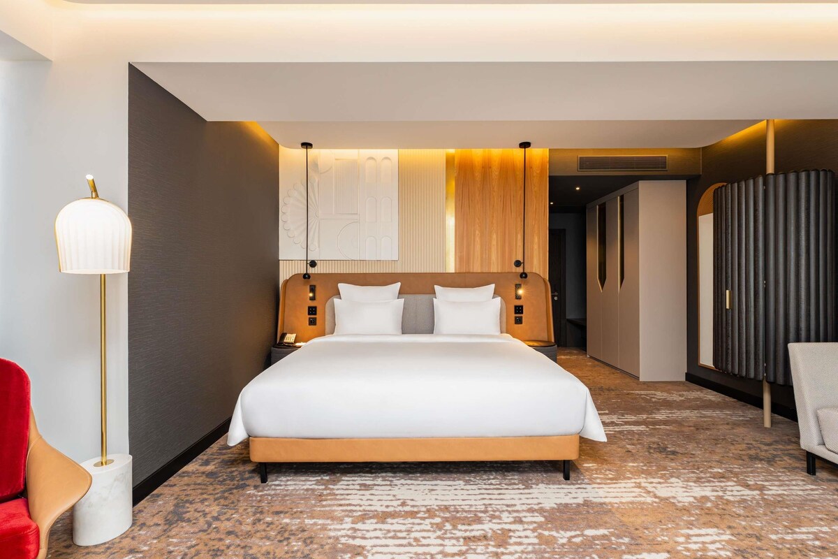 Une chambre d’hôtel avec un grand lit, une table de chevet, une lampe, une armoire et une chaise rouge.