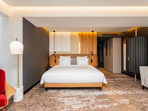 A hotel room with a large bed, a bedside table, a lamp, a wardrobe, and a red chair. - Mövenpick Yerevan (Yerevan)