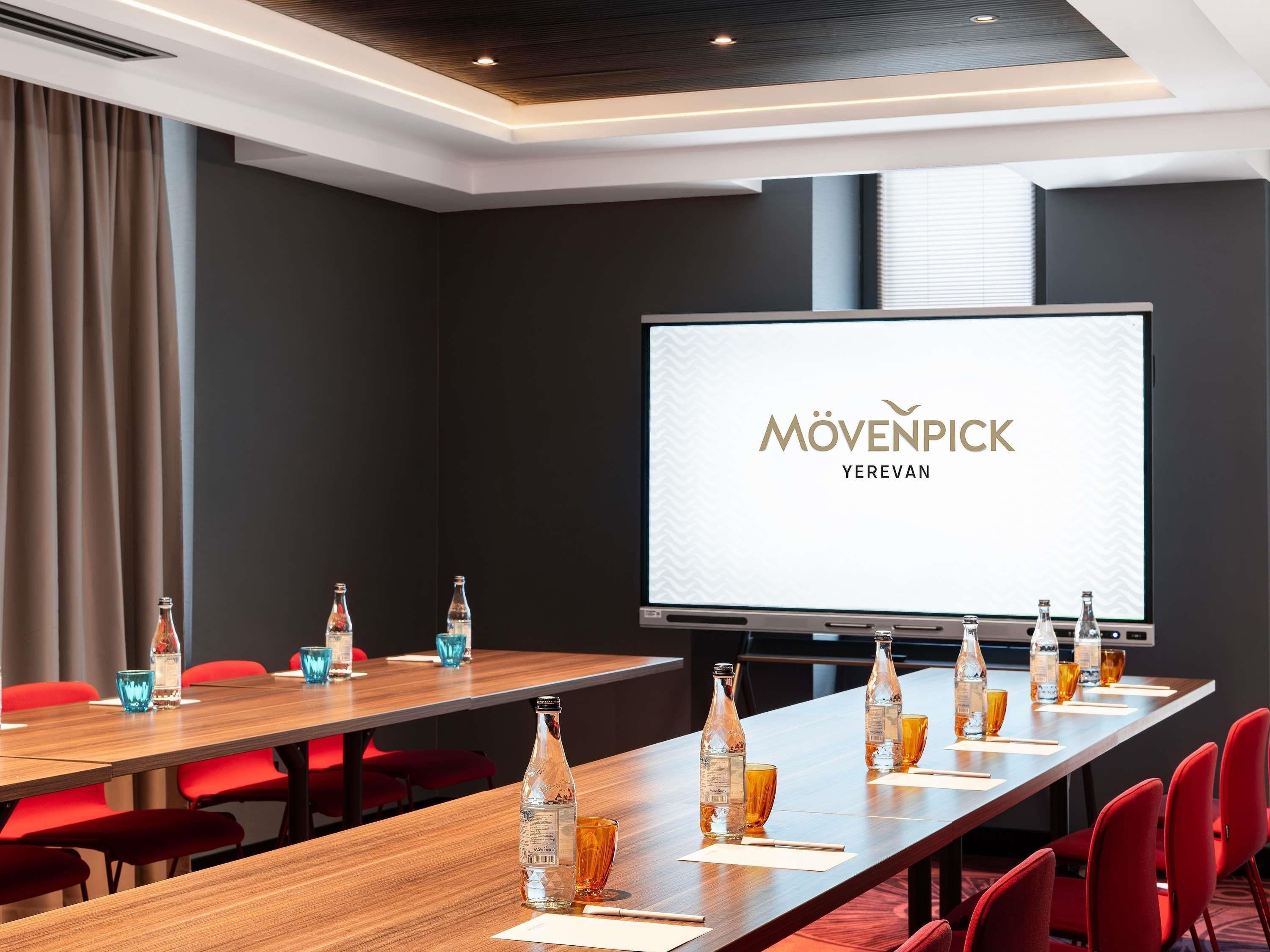Et mødelokale med et langt bord, røde stole og et skærmbillede der viser 'MÖVENPICK YEREVAN'.