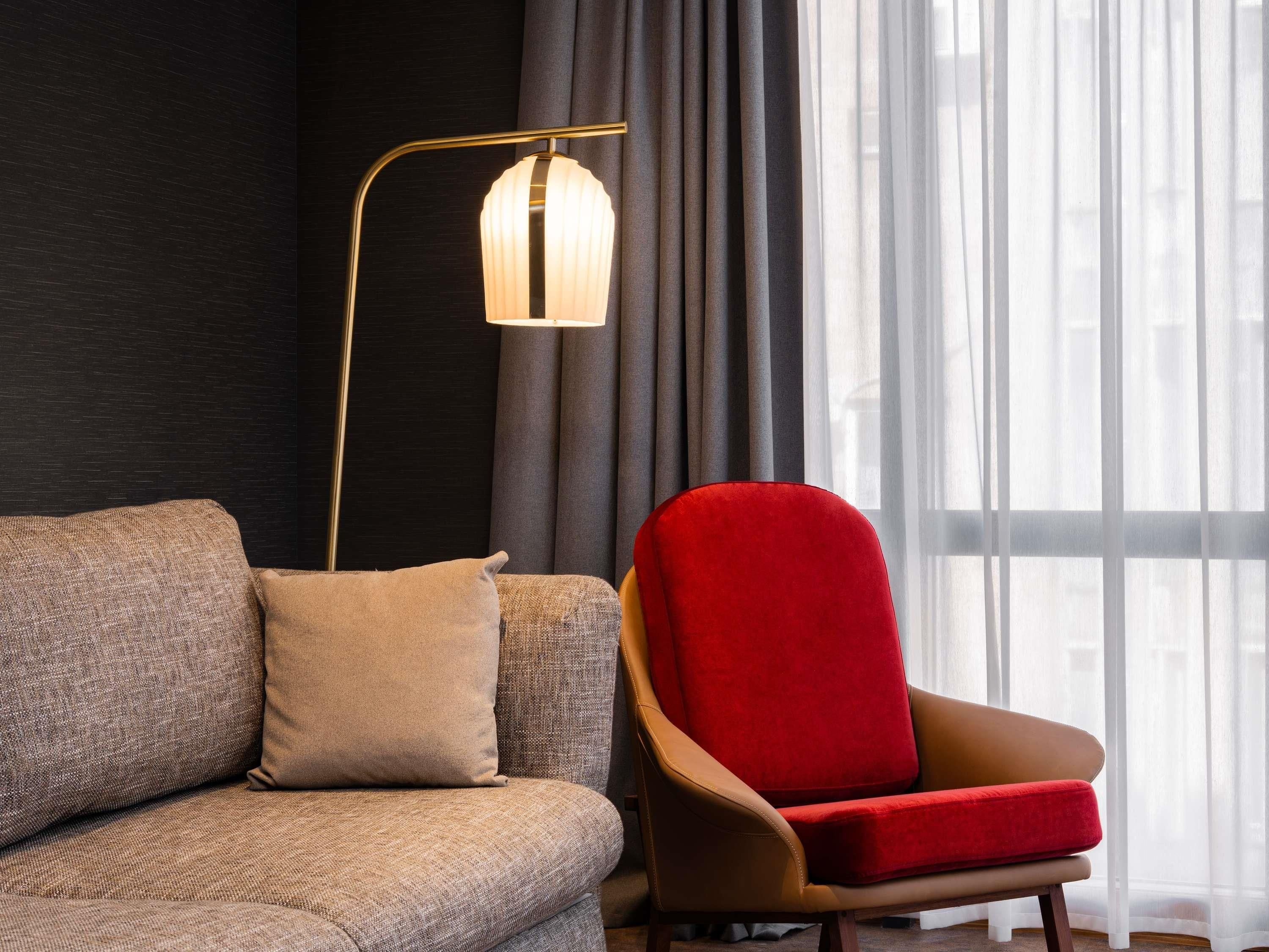 A modern living room with a sofa, a red armchair, a floor lamp, and a window with sheer curtains.
