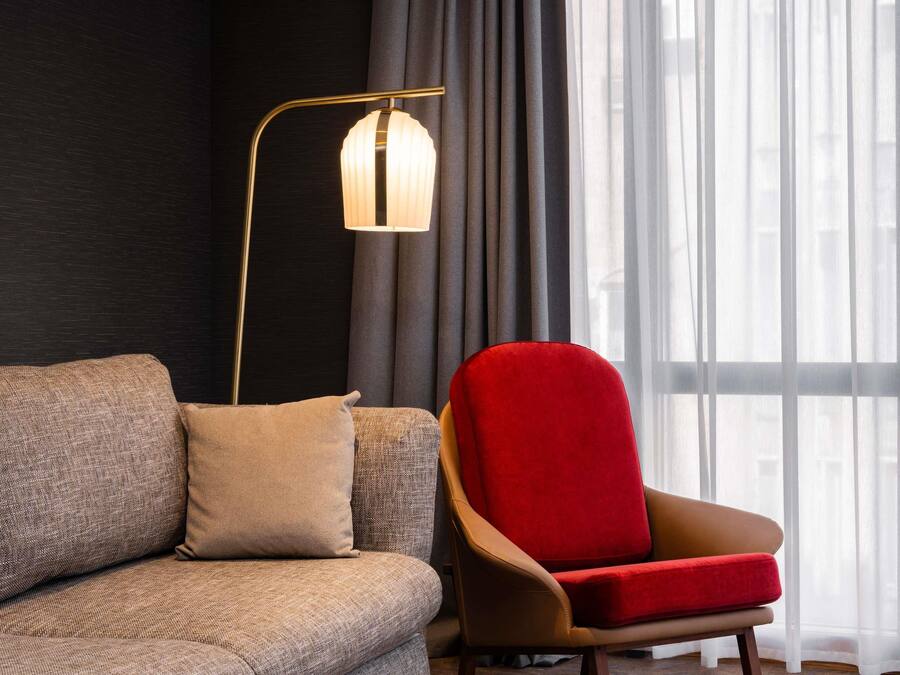 A modern living room with a sofa, a red armchair, a floor lamp, and a window with sheer curtains.