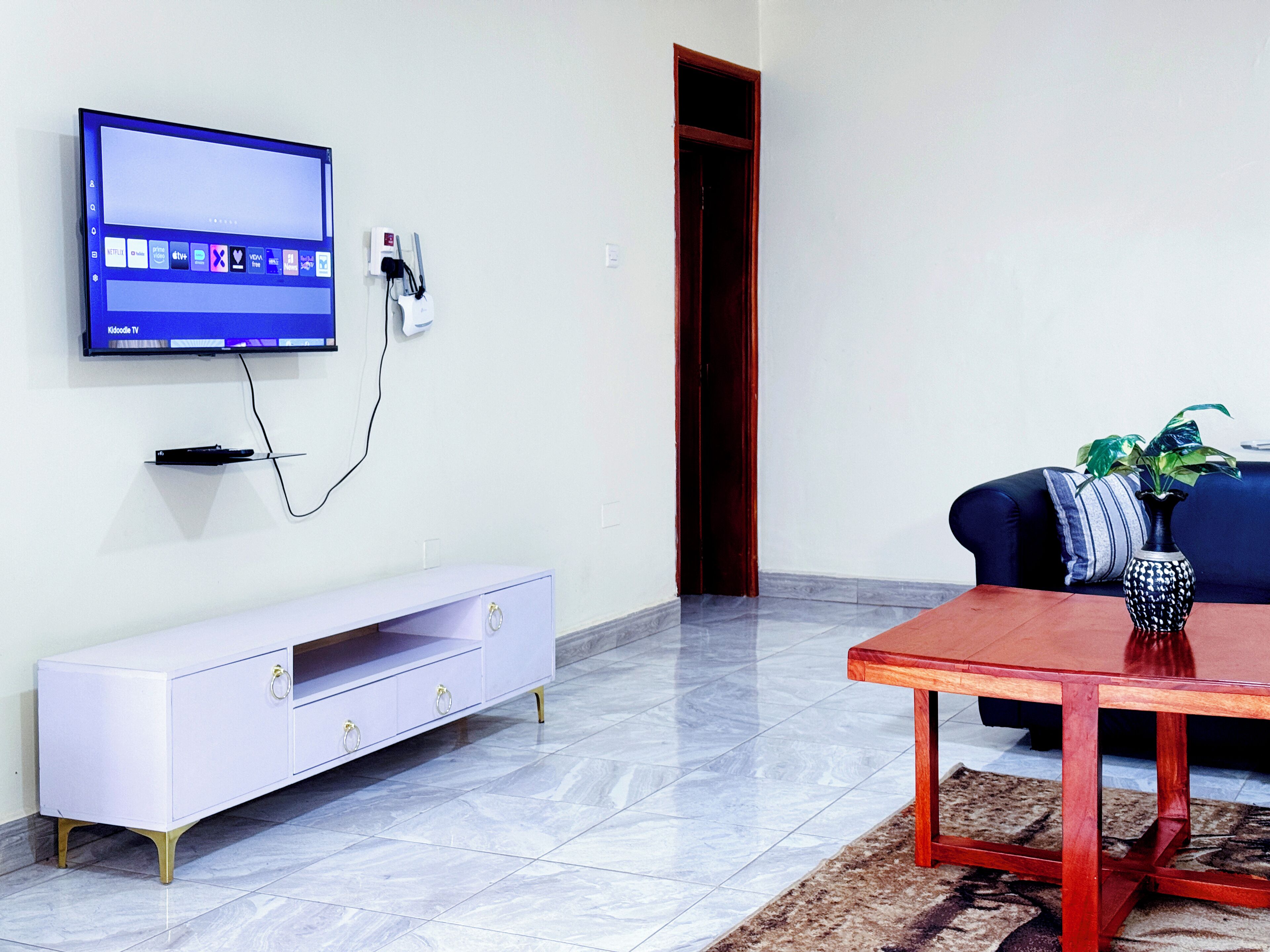 A living room with a flat-screen TV mounted on the wall, a white entertainment unit, a wooden coffee table, and a blue sofa with a plant.