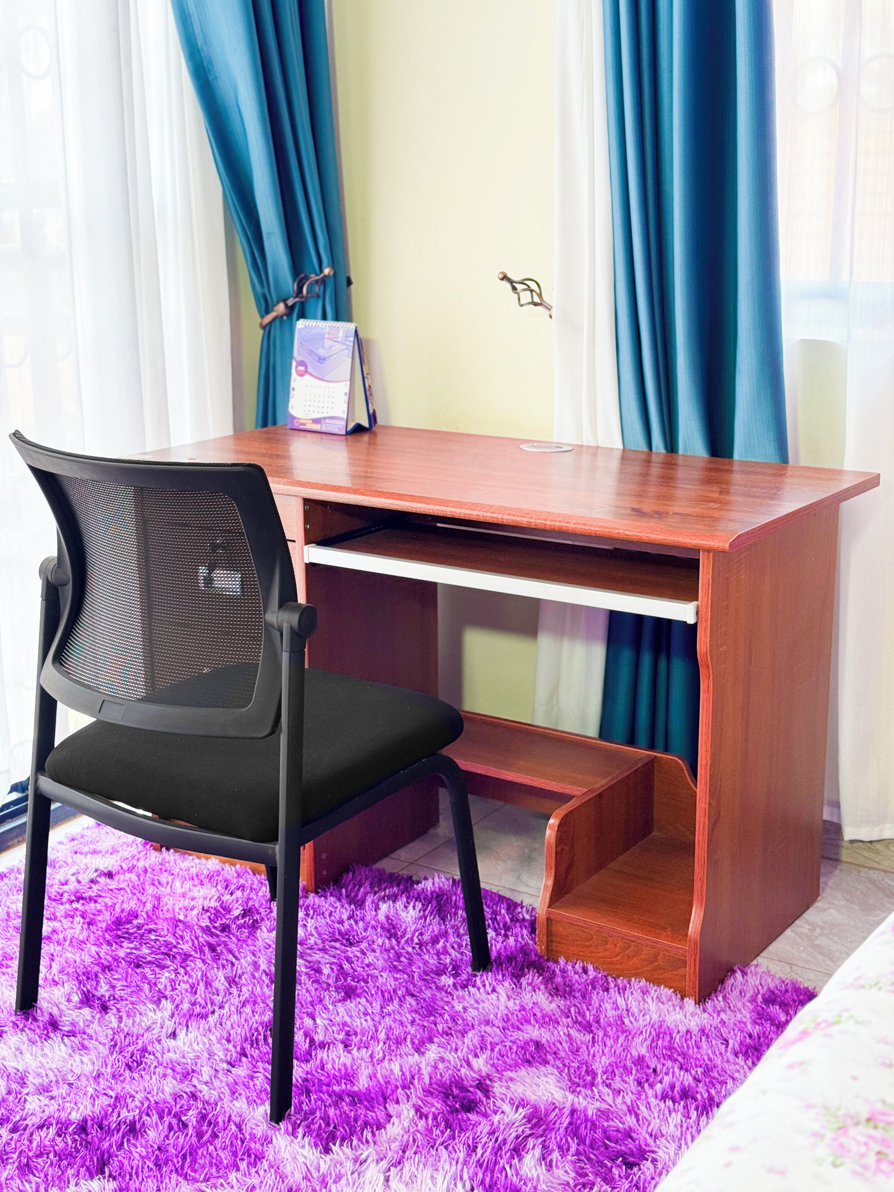 Un escritorio de madera con una silla de malla negra, un tapete mullido morado y cortinas azules al fondo.