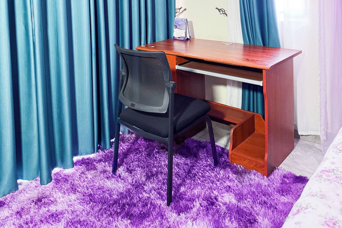 A room with a wooden desk, a black office chair, and a purple furry rug. Blue curtains are on the windows.