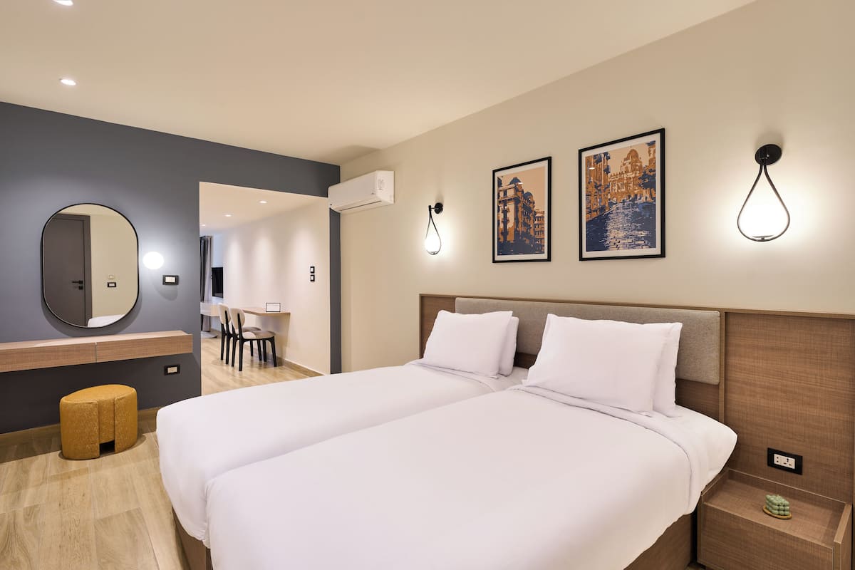 Une chambre d’hôtel avec un grand lit, un bureau, une chaise et un petit tabouret. Il y a deux lampes fixées au mur et un tableau encadré accroché au mur.