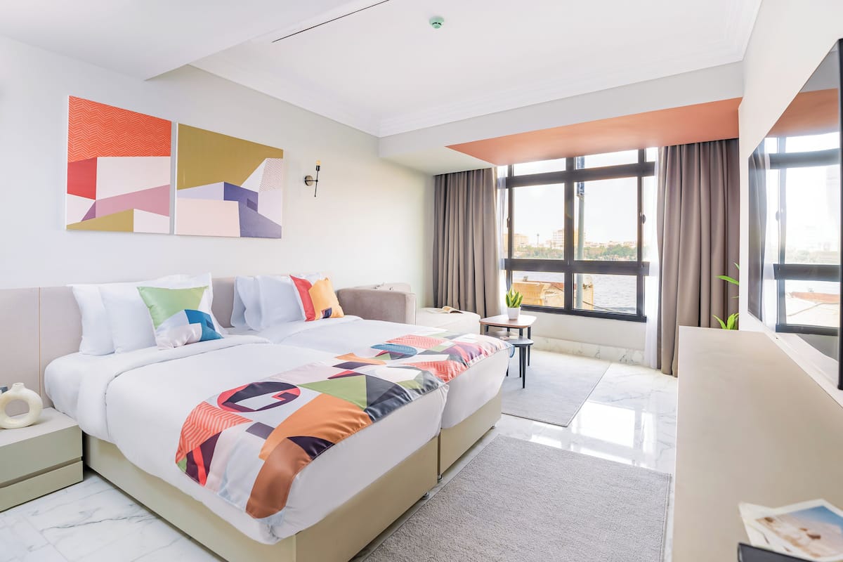 Une chambre d’hôtel moderne, dotée d’un grand lit, d’une petite table et offrant une vue sur la ville grâce à de grandes fenêtres.
