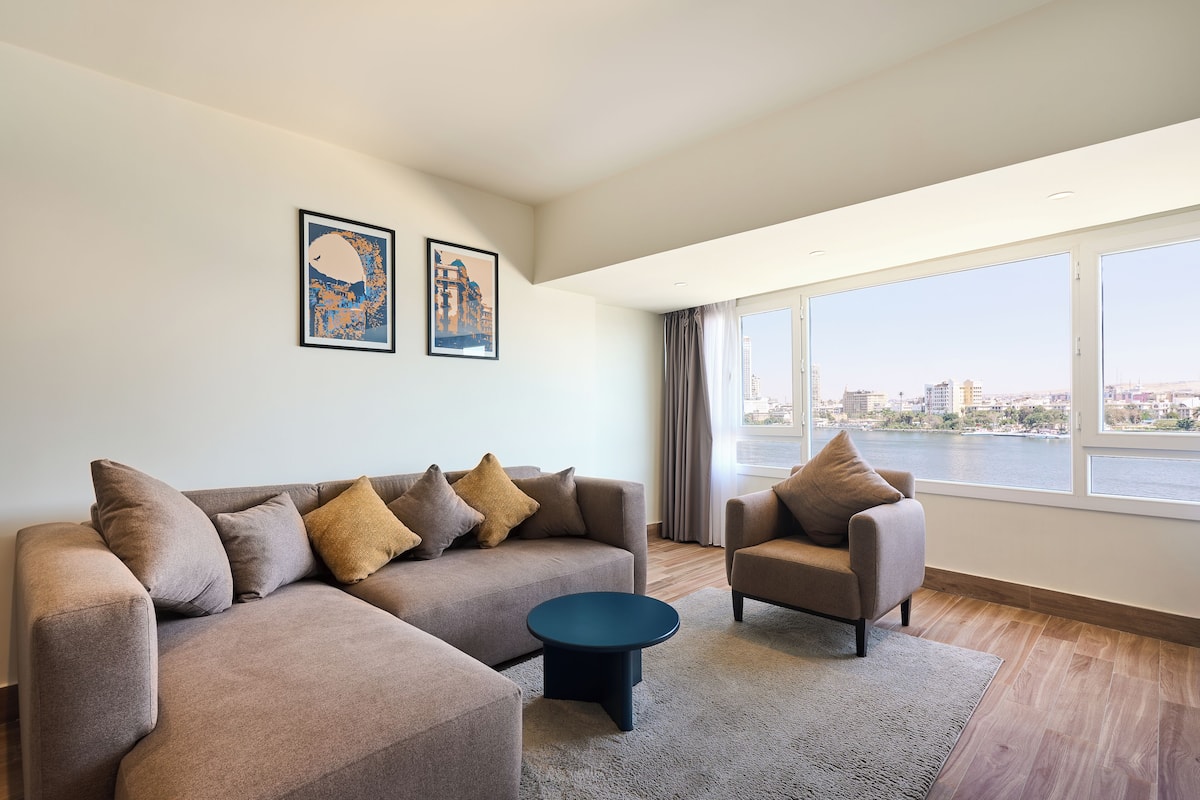 Un salon moderne avec un canapé, un fauteuil et une table basse, de grandes fenêtres offrant une vue sur la ville, et deux œuvres d’art encadrées accrochées au mur.
