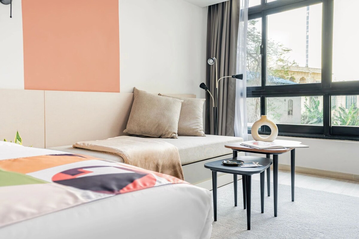 A hotel room with a bed, a small table, a lamp, and a view of the outside.