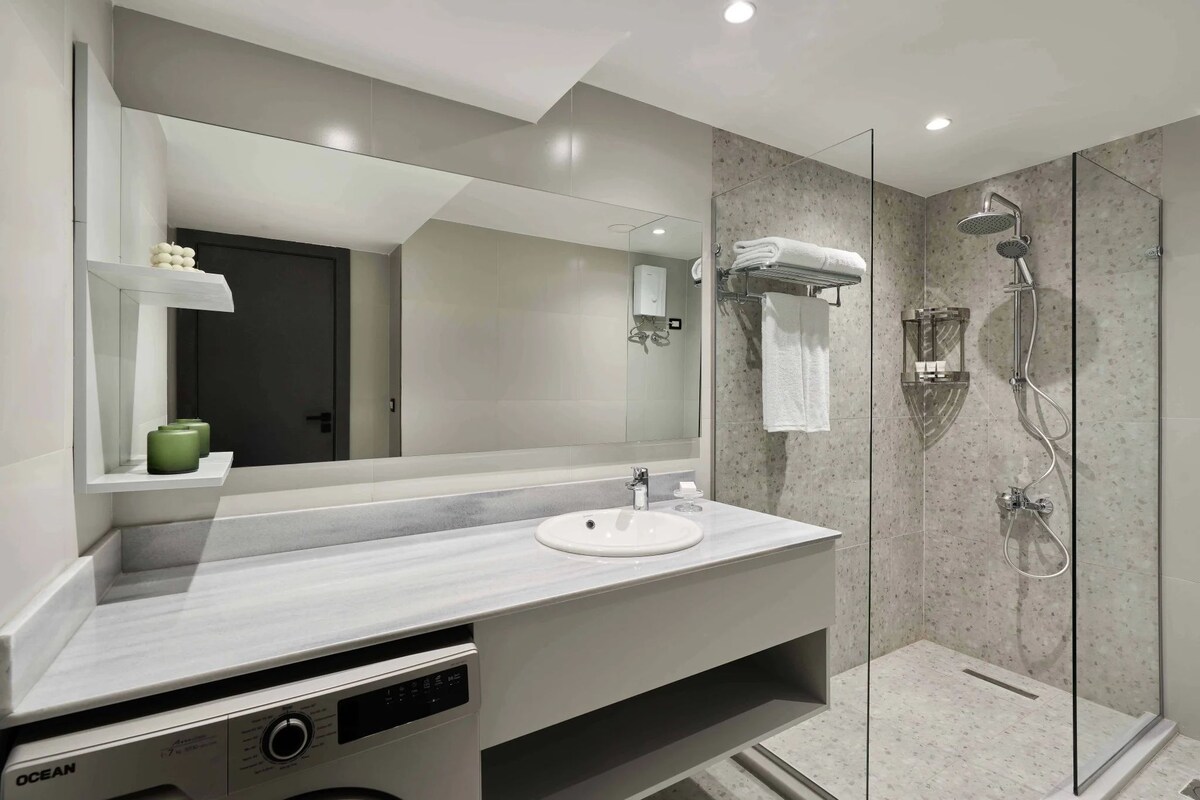 A modern bathroom with a large mirror, a sink, a shower, and a built-in washing machine.