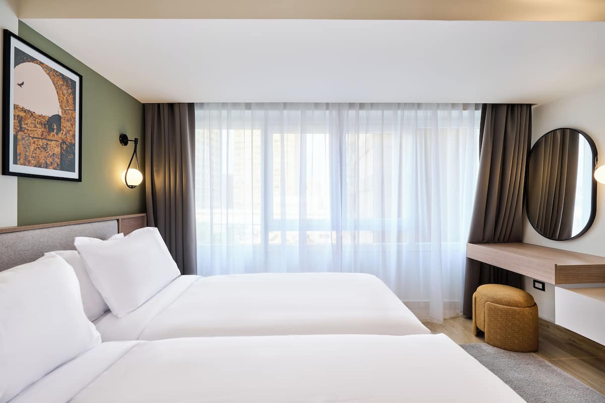 Une chambre d’hôtel avec deux lits, un bureau, une chaise, une grande fenêtre avec des rideaux légers et un miroir fixé au mur.