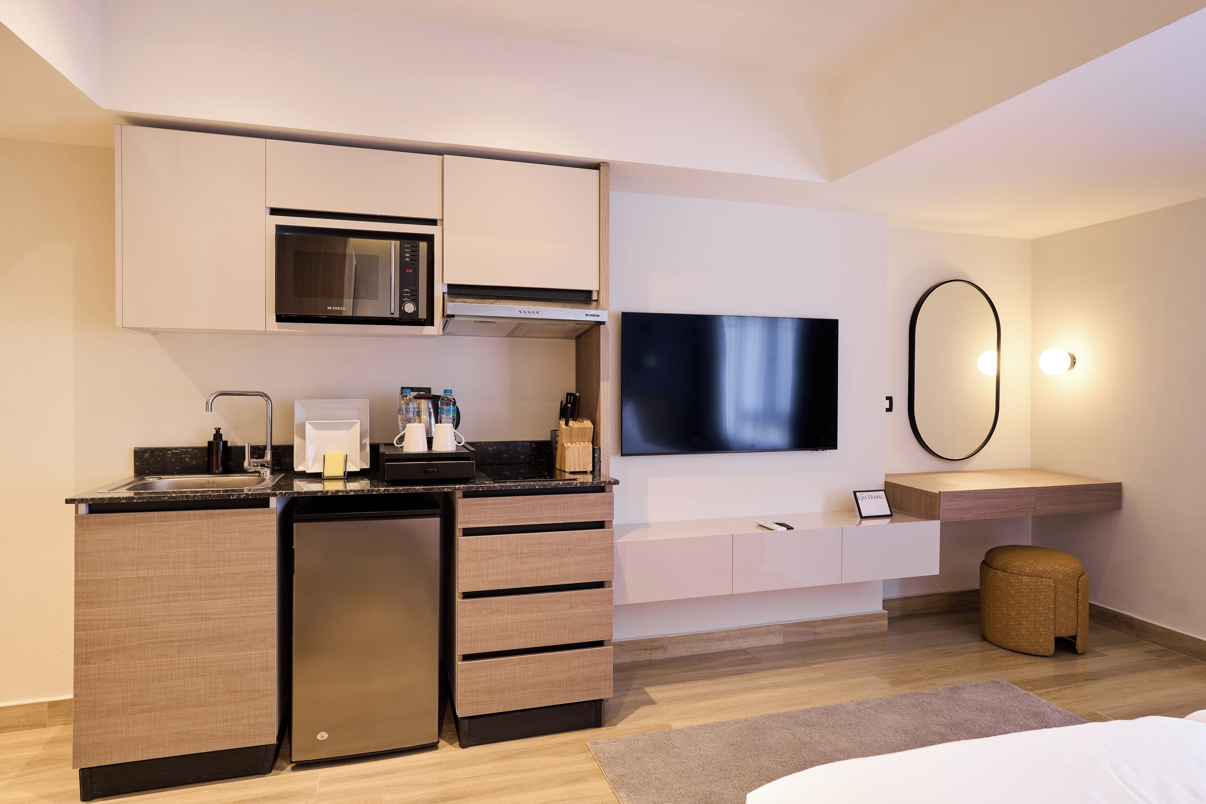 Una habitación de hotel moderna con kitchenette, televisor de pantalla plana, una pequeña zona de estar y un espejo grande.