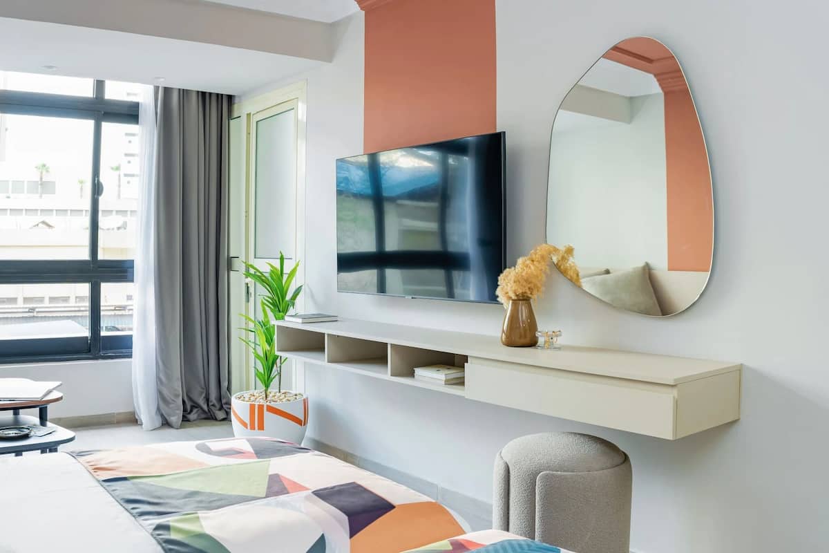 Une chambre d’hôtel moderne, dotée d’un grand lit, d’une télévision, d’un miroir, d’une plante et offrant une vue sur l’extérieur.