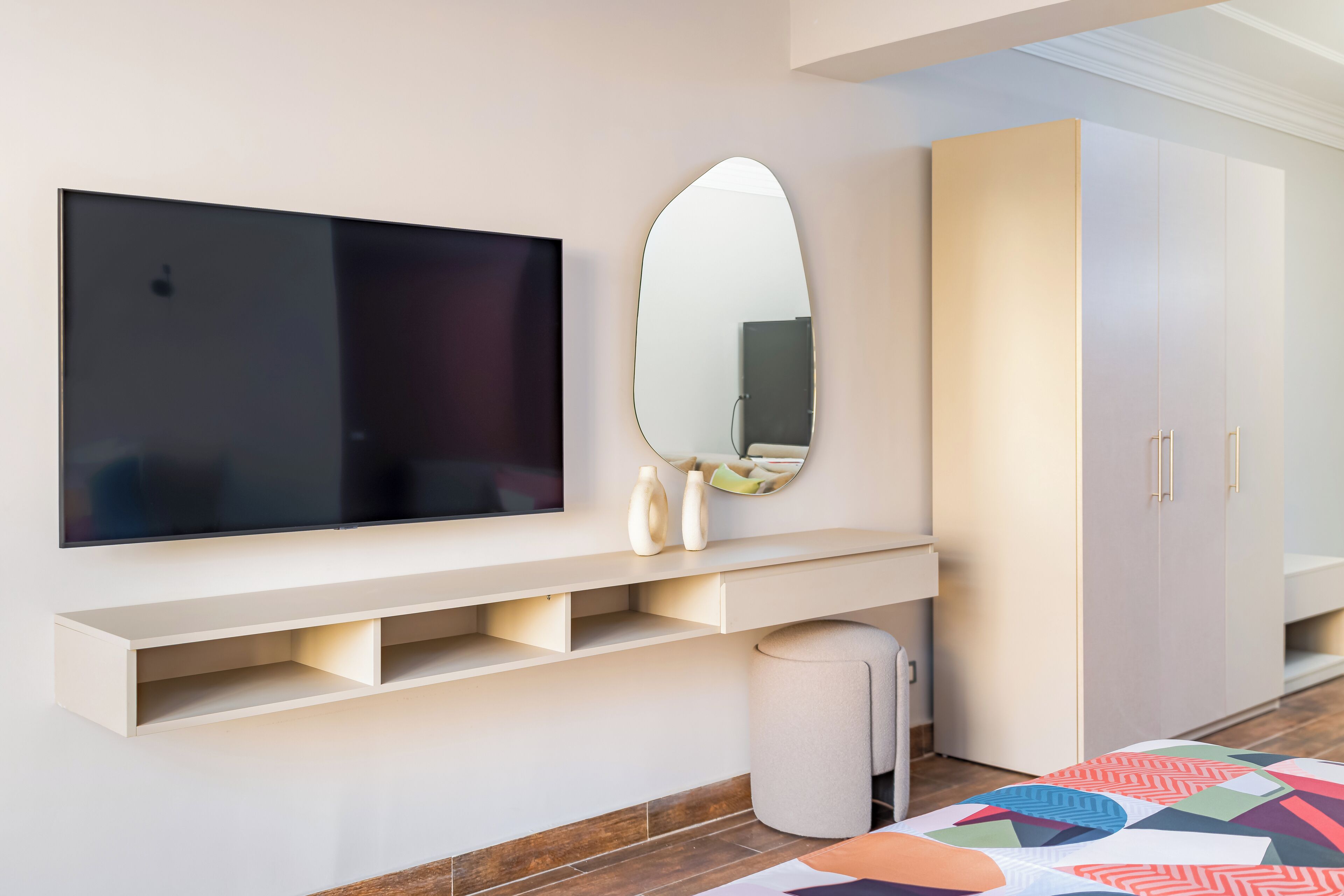 A modern bedroom with a large flat-screen TV mounted on the wall, a round mirror, a wardrobe with sliding doors, and a colorful geometric-patterned rug on the floor.