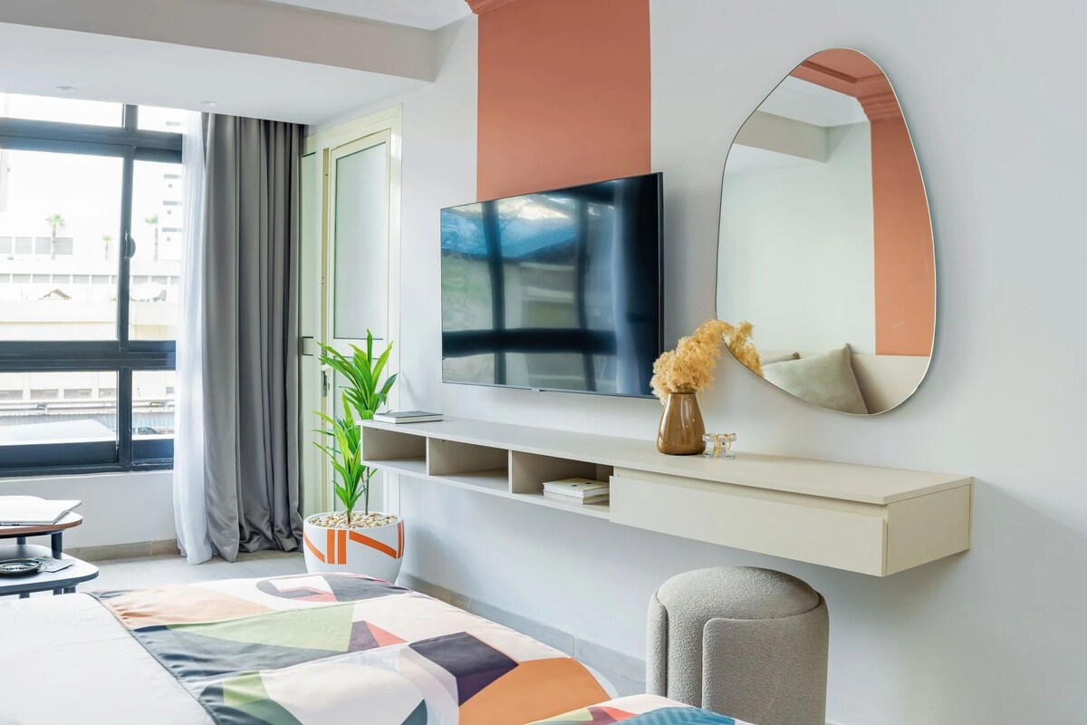 Quarto de hotel moderno com uma cama grande, televisão, espelho, planta e vista para o exterior.