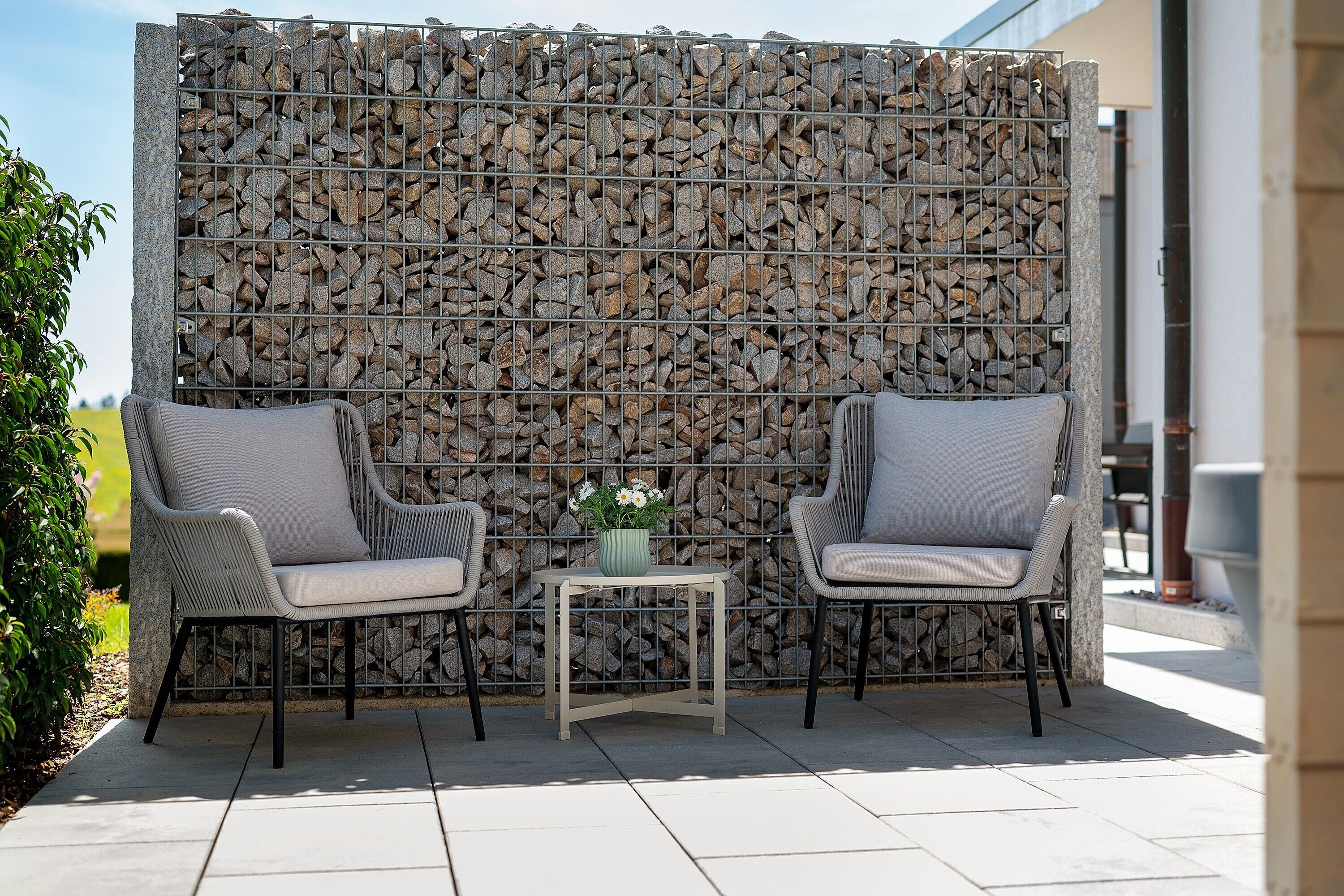 Een terras met twee rieten stoelen, een kleine tafel met een bloempot en een achtergrond van een stenen muur.