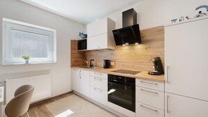 Une cuisine moderne avec des meubles blancs, un plan de travail en bois, un four encastré et une fenêtre avec des stores.