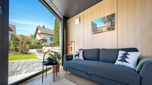 A modern living room with a grey sofa, a wooden wall panel, and a view of a house and garden. - Tiny Home 'Seewald - Tiny House' with Private Terrace, Shared Garden and Wi-Fi (Seewald)