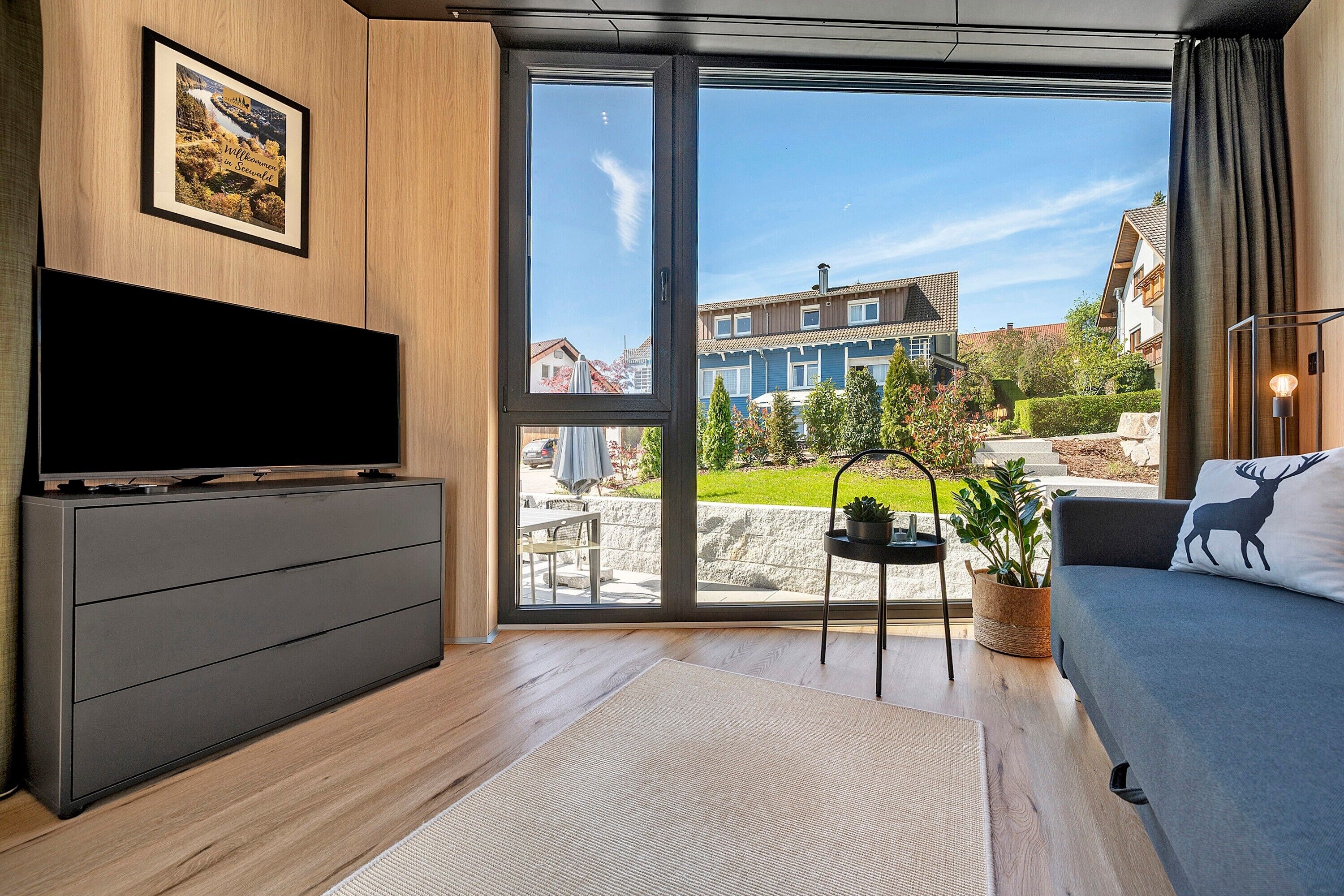 A modern living room with a flat-screen TV, a sofa with a deer pillow, a large window with a view of buildings, and a wooden cabinet.