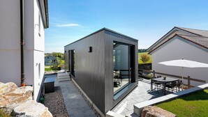 A modern building with a large glass door, a patio with outdoor furniture, and a stone pathway. - Tiny Home 'Seewald - Tiny House' with Private Terrace, Shared Garden and Wi-Fi (Seewald)