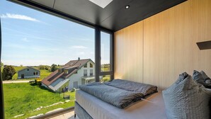 Slaapkamer met een groot raam, een bed en uitzicht op een huis en een groen landschap.