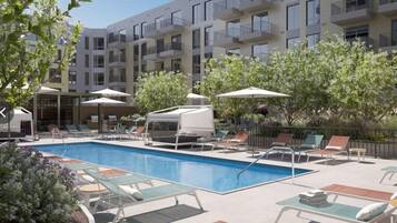 Una piscina con sillones y sombrillas, rodeada por un edificio de varios pisos con balcones.