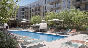 Une piscine entourée de chaises longues et de parasols, elle-même bordée d’un immeuble à plusieurs étages doté de balcons.