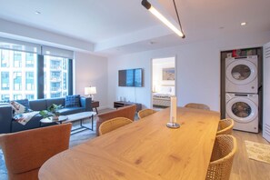 Un salon moderne avec une table à manger en bois, des sièges confortables, une grande fenêtre et un coin buanderie équipé d’un lave-linge.