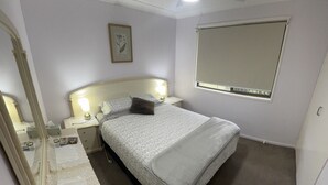 A bedroom with a bed, a bedside table, a nightstand, a ceiling fan, a window with a blind, and a framed picture on the wall. - Quiet cozy room by the beach R1 (Brighton)