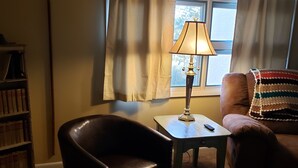 A cozy living room with a leather armchair, a small table with a lamp, a bookshelf, and a window with curtains.