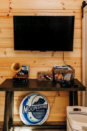 Una pared de madera con un televisor montado, un tocadiscos antiguo, una mesita con una cesta y un letrero circular en el suelo.