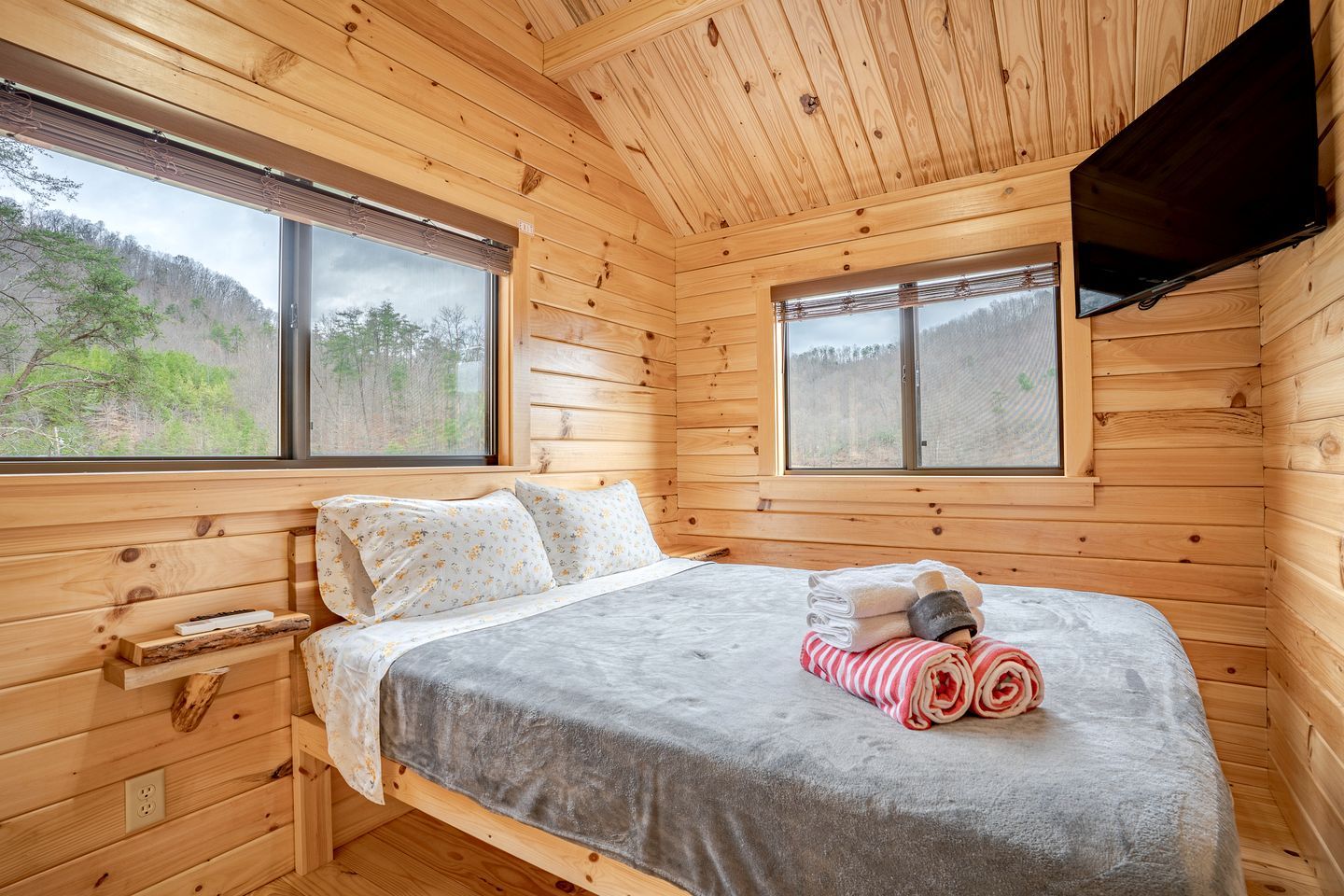 Un dormitorio de cabaña de madera con una cama, dos ventanas, un televisor y vistas a un bosque.
