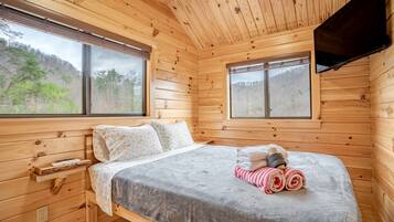Un dormitorio de cabaña de madera con una cama, dos ventanas, un televisor y vistas a un bosque.