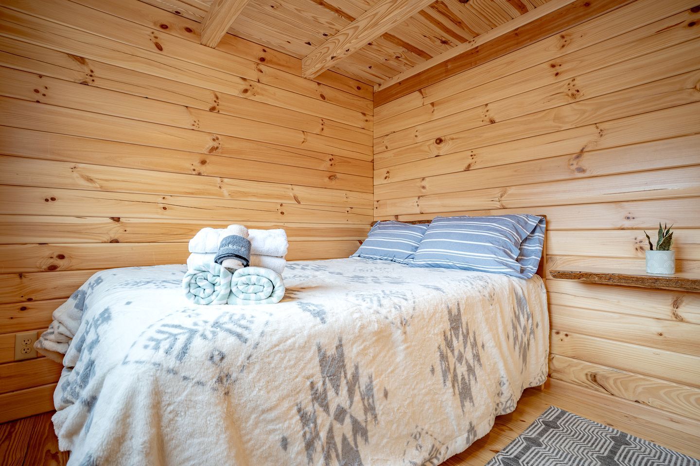 A wooden cabin room with a bed, pillows, a rolled-up towel, a small shelf with a plant, and a rug.