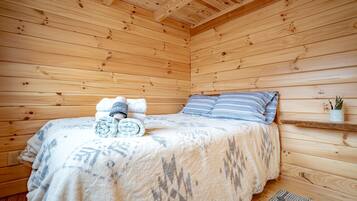 A wooden cabin room with a bed, pillows, a rolled-up towel, a small shelf with a plant, and a rug.