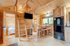 Ein Holzblockhaus-Innenraum mit einem Bett, einem an der Wand befestigten Fernseher, einer Küchenzeile inklusive Kühlschrank und einem Esstisch mit Hocker.