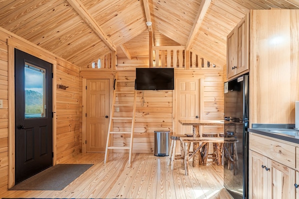 A wooden cabin interior with a kitchen area, a dining table, a ladder, and a door with a view.