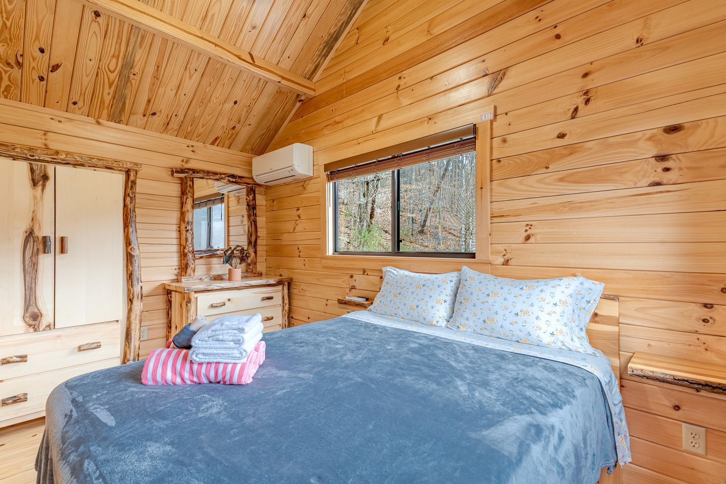 Un dormitorio de cabaña de madera con cama, cómoda y ventana con vistas a árboles.
