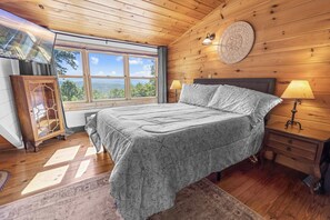 Quarto com painéis de madeira, cama, duas luminárias de cabeceira, mesa de apoio, detalhe decorativo na parede e janela com vista para o exterior.