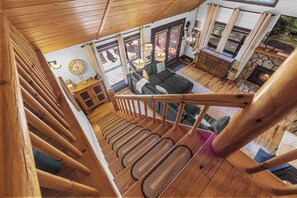 Uma escada de madeira leva a uma sala de estar com sofá, lareira e grandes janelas.