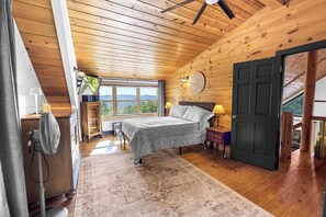 Une chambre à coucher avec un lit, des tables de chevet, un ventilateur, une fenêtre donnant sur un beau paysage et une porte.