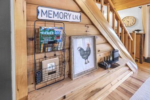 Un escalier en bois avec une pancarte indiquant « MEMORY LN. », un tableau encadré représentant un coq et un présentoir en métal contenant des magazines et une télécommande.