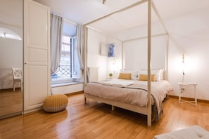 A bedroom with a four-poster bed, a window with curtains, a radiator, and a small bedside table.