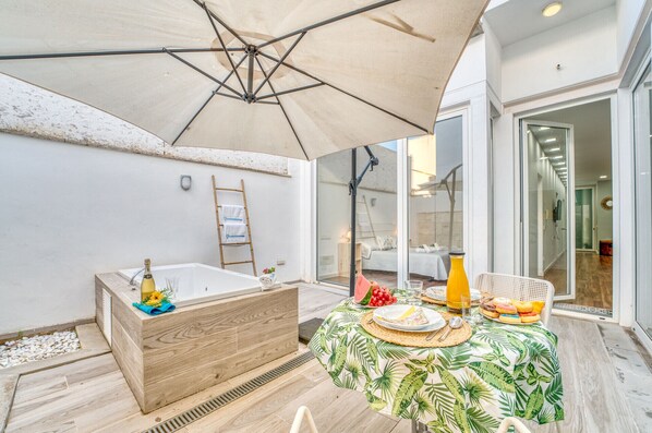 A patio area with a wooden bathtub, a table set for breakfast, and a large umbrella providing shade.