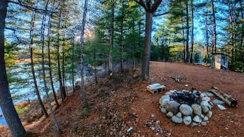 A forested campsite with a fire pit, a small shed, and a dirt path leading to a body of water.