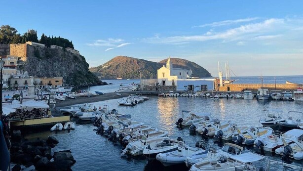A coastal marina with boats docked, a church, and buildings on a hillside.