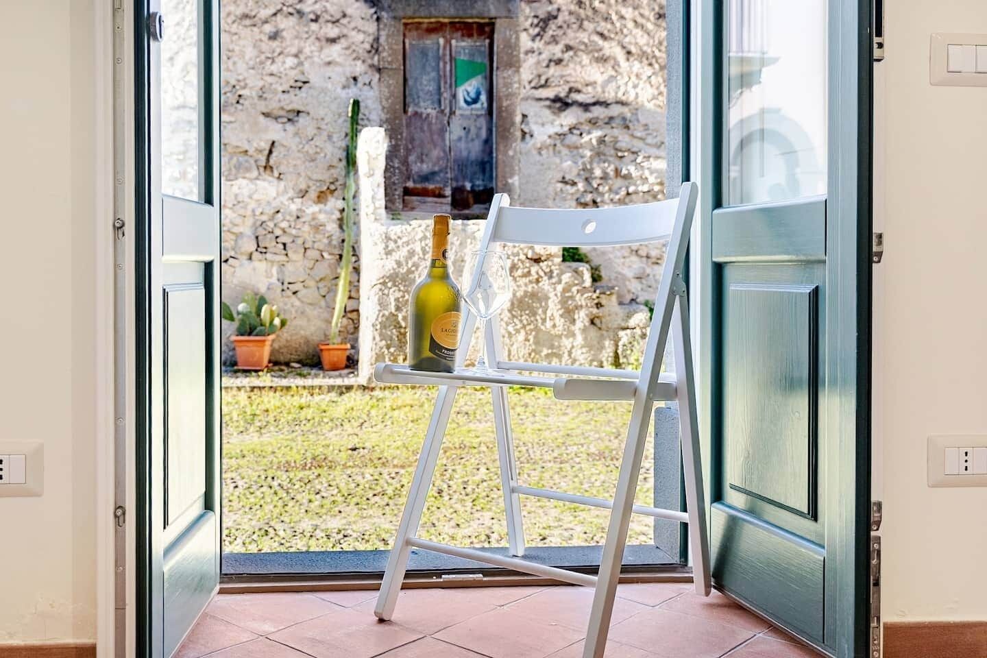 A room with a white chair, a ladder, a bottle of wine, and a view of an outdoor area with a stone wall and potted plants.