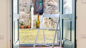 A room with a white chair, a ladder, a bottle of wine, and a view of an outdoor area with a stone wall and potted plants.