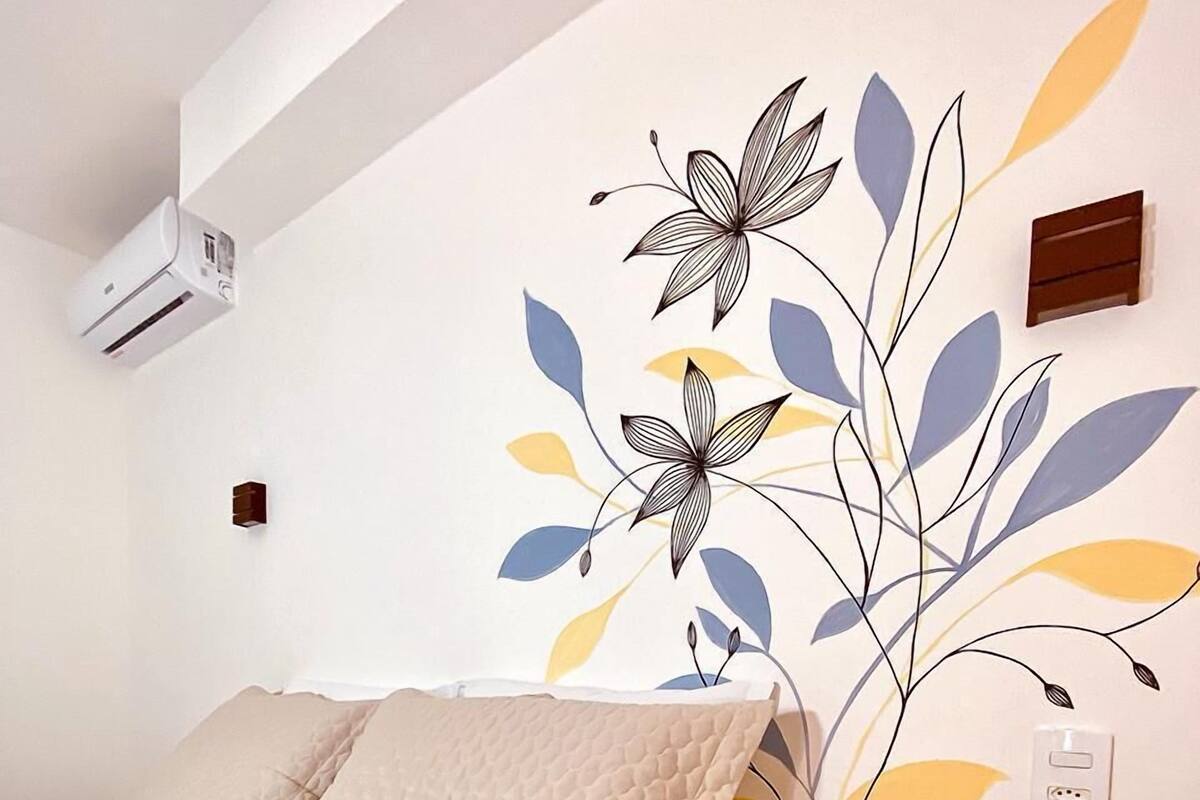 A bedroom with a bed, a wall-mounted air conditioner, a bedside table with a remote control, and a wall decoration.